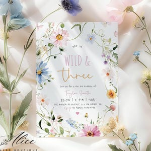 Może przedstawiać: Kwiatowe zaproszenie z napisem "she is WILD & Three" w złotym skrypcie. Zaproszenie otoczone jest akwarelowymi kwiatami w odcieniach różu, błękitu i żółci. Zaproszenie na 3. urodziny.