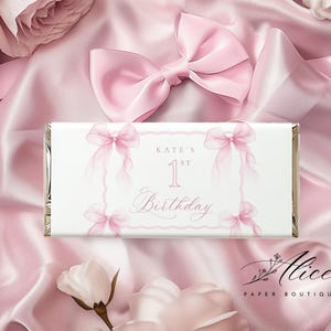 Peut inclure: Emballage de barre chocolatée blanche avec des nœuds roses et le texte "Kate's 1st Birthday" sur un fond de satin rose avec des roses roses et un nœud. Le logo Alice Paper Boutique est en bas à droite.