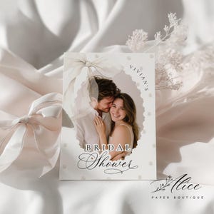 K&ouml;nnte beinhalten: Eine Einladungskarte zur Brautparty mit einem sich umarmenden Paar in einem ovalen Rahmen. Die Karte hat eine weiche, neutrale Farbpalette mit den Worten "Bridal Shower" in eleganter Schrift. Die Karte ist mit Perlen und einer Schleife verziert.