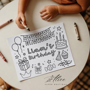 May include: A white rectangular coloring sheet featuring black line art of a birthday cake, balloons, gifts, a crown, and candy. The text reads "HAPPY BIRTHDAY" on a banner and "Liam's Birthday" in the center. Several crayons are placed nearby.