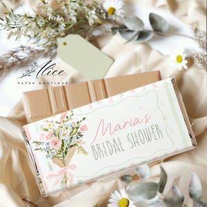 May include: A chocolate bar with a floral design and the text "Maria's Bridal Shower." The wrapper features a bouquet of flowers in pink, blue, and white, tied with a pink ribbon. A second chocolate bar is visible in the background.