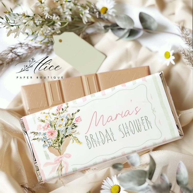 May include: A chocolate bar with a floral design and the text "Maria's Bridal Shower." The wrapper features a bouquet of flowers in pink, blue, and white, tied with a pink ribbon. A second chocolate bar is visible in the background.