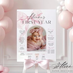 May include: A pink and white first year milestone sign featuring a baby in a pink bow. The sign displays the baby's weight, teeth, and height in inches. The sign is decorated with a pink bow. The baby is 66 cm tall.