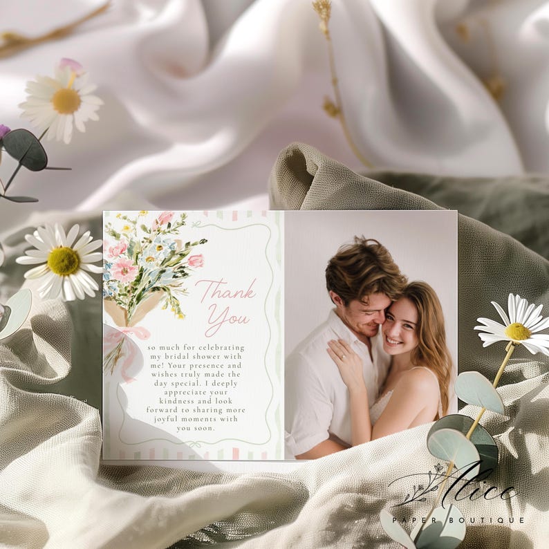 May include: A bridal shower thank you card with a floral bouquet illustration and the words "Thank You." The card also features a photograph of a couple. The card is set against a backdrop of white and green fabric.
