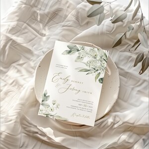 May include: A white wedding invitation with green foliage and white flowers. The invitation reads "Together with their families, Emily Bennet & Jeremy Smith, Jolly invite you to celebrate their wedding, Sep 15, 2023, Ceremony to begin at 3 PM, Villa Dante, Via Nino 13, Varese, Reception to follow."
