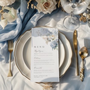 Może przedstawiać: Biała karta menu ślubnego z kwiatowym wzorem z niebieskich hortensji i białych r&oacute;ż. Menu zawiera przystawki, dania gł&oacute;wne, desery oraz imiona Mary i William z datą 21 września 2023.