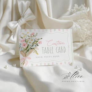 May include: A custom table card with a floral bouquet design. The card features the text "Custom Table Card" and "Your Texts Here". The bouquet includes pink, blue, and white flowers tied with a pink ribbon. The card is set against a white background.