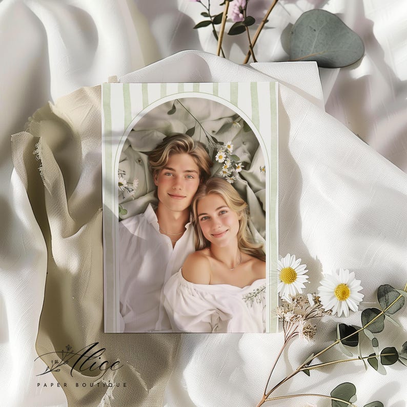 May include: A greeting card featuring a couple lying down, framed by a green and white striped arch. The card is surrounded by white fabric, flowers, and greenery. The card has the text "Alice Paper Boutique".