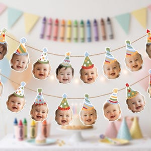 May include: A decorative party banner featuring a series of cutouts of smiling infants wearing colorful, patterned party hats. The cutouts are suspended from a thin, natural-colored twine string, arranged in two horizontal rows for display.