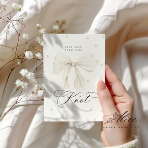 K&ouml;nnte beinhalten: Eine cremefarbene Karte mit einem Aquarell-Bogen und den Worten "Lily Has Tied The Knot". Die Karte ist mit kleinen Perlenakzenten verziert und wird von einer Hand gehalten. Das Logo von Alice Paper Boutique befindet sich unten rechts.