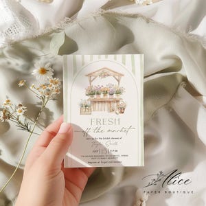 May include: A floral-themed invitation card with a watercolor illustration of a flower stand. The card features the text "FRESH off the market" and details for a bridal shower, including the date, time, and location. The card is held against a soft, textured background.