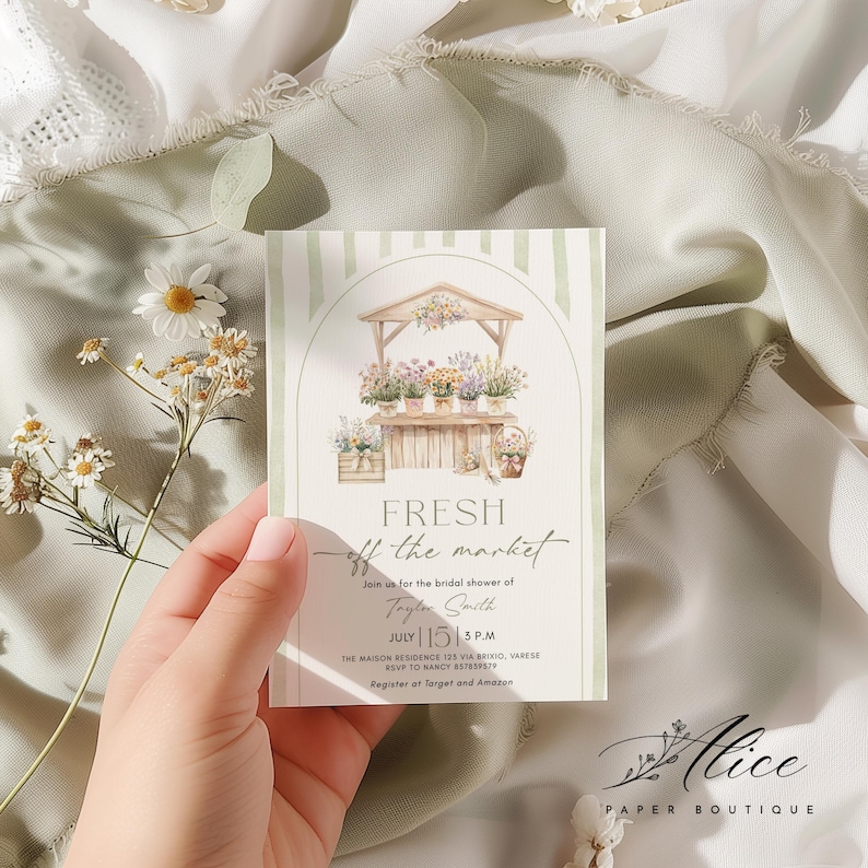 May include: A floral-themed invitation card with a watercolor illustration of a flower stand. The card features the text "FRESH off the market" and details for a bridal shower, including the date, time, and location. The card is held against a soft, textured background.