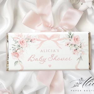 Puede incluir: Envoltorio blanco de barra de caramelo con el texto "ALICIA'S Baby Shower" en escritura rosa, decorado con rosas rosas y un lazo rosa. El envoltorio está sobre un fondo de tela de satén blanco y una cinta rosa.