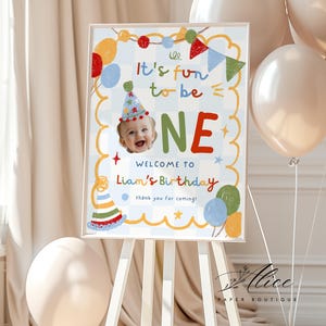 May include: A rectangular birthday sign featuring a light blue checkered background, colorful pennant banners, and balloons. The text reads It's fun to be ONE, WELCOME TO Liam's Birthday, and thank you for coming. A photo of a baby wearing a party hat is included.