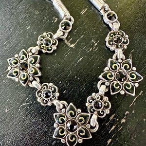 May include: Silver necklace with a floral design, black and gold detailing. The necklace features flower-shaped pendants with black central stones. The chain is silver, and the piece is set against a dark background. The necklace is a piece of jewellery.