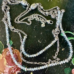 May include: A multi-strand necklace with silver-toned chains and light blue crystal beads. The necklace features decorative silver accents and a lobster clasp closure. The design includes multiple layers of chains and beaded strands.