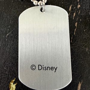 May include: A rectangular silver-toned metal dog tag pendant with a brushed finish, featuring the text &copy; Disney printed in black near the bottom. The pendant is attached to a silver-toned ball chain necklace through a small hole at the top.