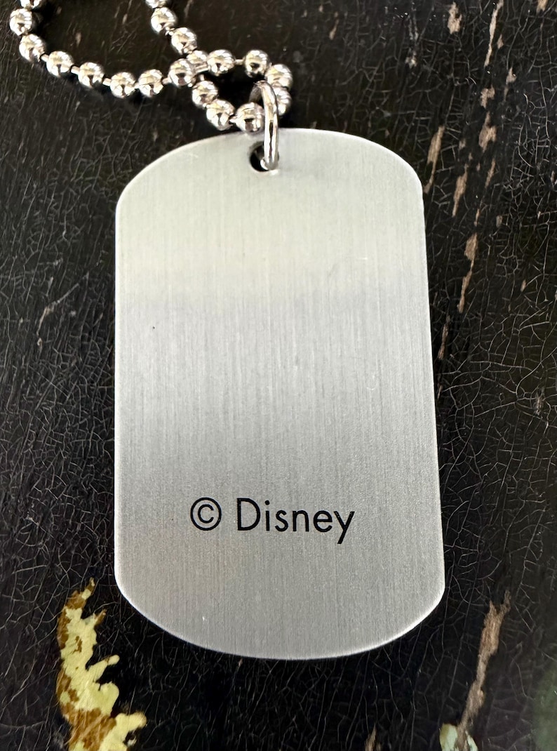 May include: A rectangular silver-toned metal dog tag pendant with a brushed finish, featuring the text &copy; Disney printed in black near the bottom. The pendant is attached to a silver-toned ball chain necklace through a small hole at the top.