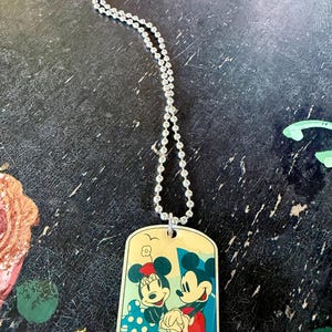 May include: A silver-toned ball chain necklace featuring a rectangular dog tag pendant. The pendant displays a colorful illustration of Mickey and Minnie Mouse holding hands, set against a light yellow background with a small bird graphic above.