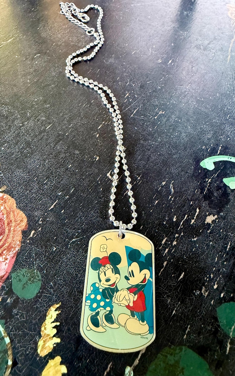 May include: A silver-toned ball chain necklace featuring a rectangular dog tag pendant. The pendant displays a colorful illustration of Mickey and Minnie Mouse holding hands, set against a light yellow background with a small bird graphic above.