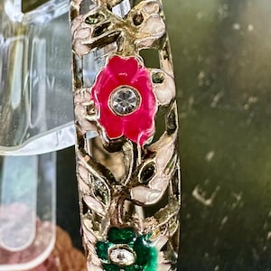May include: A gold-toned metal bracelet with a floral design. The bracelet features cut-out leaves and flowers in pink and green, each with a central crystal. The bracelet is a decorative accessory.