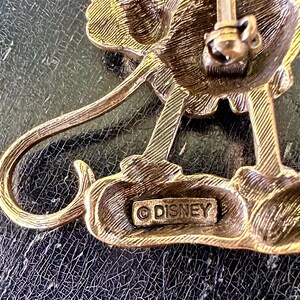 May include: Vintage gold-toned metal Disney brooch. The brooch has a detailed design and a pin closure. The word "DISNEY" is stamped on the back. The brooch is on a dark, textured surface.