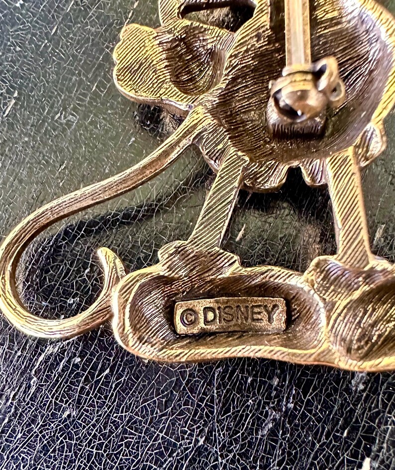 May include: Vintage gold-toned metal Disney brooch. The brooch has a detailed design and a pin closure. The word "DISNEY" is stamped on the back. The brooch is on a dark, textured surface.