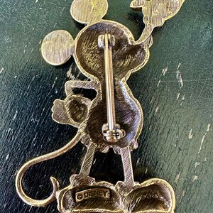 May include: Vintage gold-toned Mickey Mouse brooch. The pin features Mickey Mouse holding a flag. The back of the pin has a clasp and the word "DISNEY" is stamped on the bottom. The brooch is made of a gold-coloured metal.