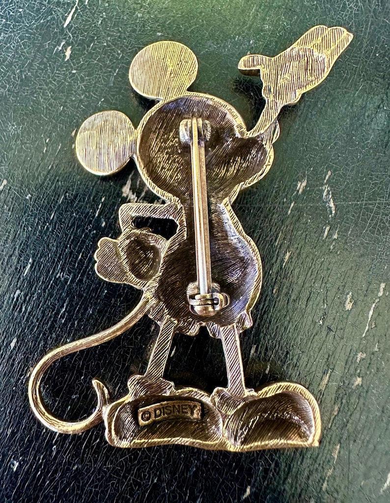 May include: Vintage gold-toned Mickey Mouse brooch. The pin features Mickey Mouse holding a flag. The back of the pin has a clasp and the word "DISNEY" is stamped on the bottom. The brooch is made of a gold-coloured metal.