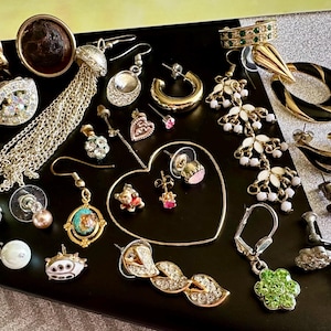 May include: A collection of various earrings in gold, silver, and rhinestone. Styles include hoops, studs, and dangling designs. Some earrings feature pearls, gemstones, and unique shapes, such as hearts and flowers. The earrings are displayed on a black surface.