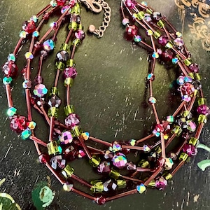 May include: A multi-strand necklace featuring a variety of beads in shades of red, purple, and green. The beads are strung on thin, reddish-brown cords and secured with a silver-colored clasp. The necklace has an iridescent quality.