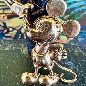 May include: A gold-coloured Mickey Mouse figurine in a standing pose, with one arm raised and the other holding a small object. The figurine has large round ears and is set against a dark background.