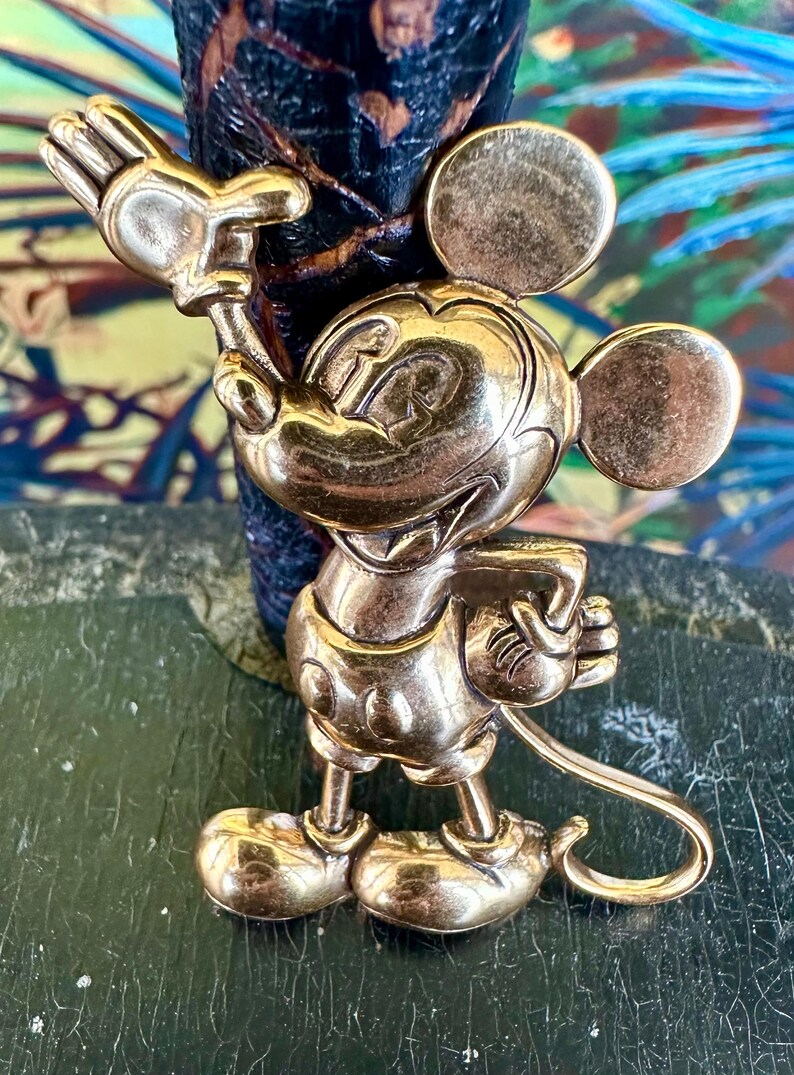 May include: A gold-coloured Mickey Mouse figurine in a standing pose, with one arm raised and the other holding a small object. The figurine has large round ears and is set against a dark background.