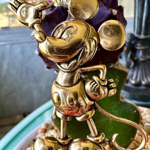 May include: A gold-coloured Mickey Mouse figurine in a cheerful pose. The figurine has rounded features and is standing on a cactus. The background includes a window and a decorative plant.