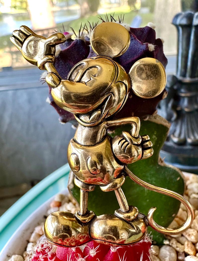 May include: A gold-coloured Mickey Mouse figurine in a cheerful pose. The figurine has rounded features and is standing on a cactus. The background includes a window and a decorative plant.
