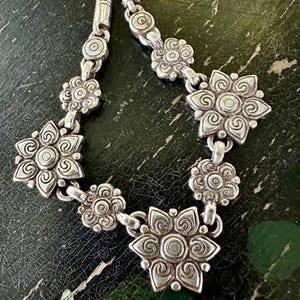 May include: A silver necklace with a floral design. The necklace features multiple flower-shaped pendants connected by small links. The flowers have detailed petal patterns and a central circular design. The chain is visible at the top, with a woven texture.