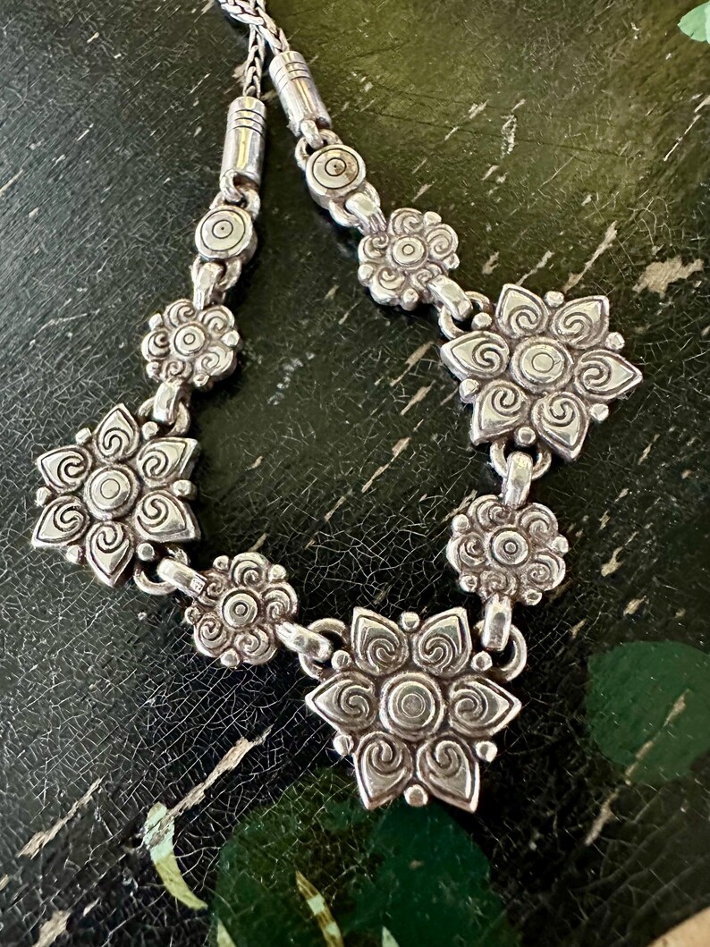 May include: A silver necklace with a floral design. The necklace features multiple flower-shaped pendants connected by small links. The flowers have detailed petal patterns and a central circular design. The chain is visible at the top, with a woven texture.
