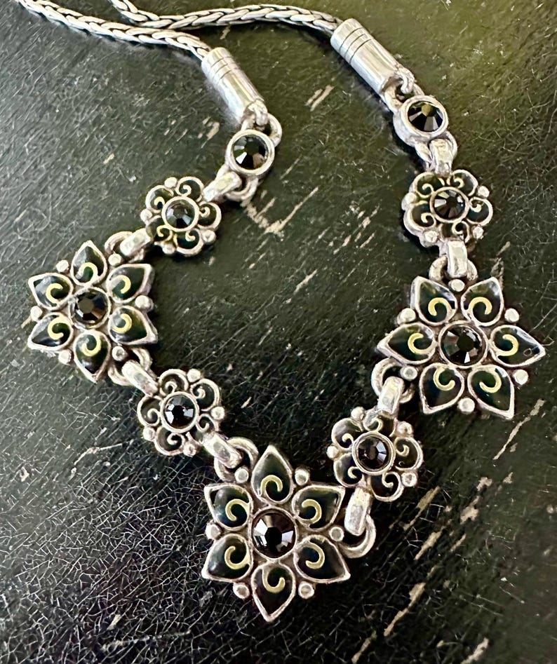 May include: Silver necklace with a floral design, black and gold detailing. The necklace features flower-shaped pendants with black central stones. The chain is silver, and the piece is set against a dark background. The necklace is a piece of jewellery.