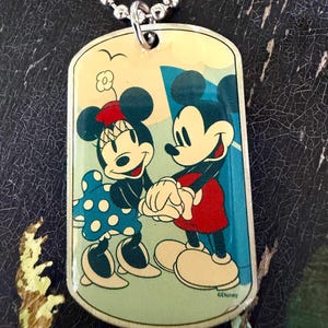 May include: A rectangular metal dog tag pendant on a silver ball chain. The pendant features a colorful graphic of Mickey and Minnie Mouse holding hands. Minnie wears a blue polka dot dress and red hat, while Mickey wears red shorts. The text Disney is visible.