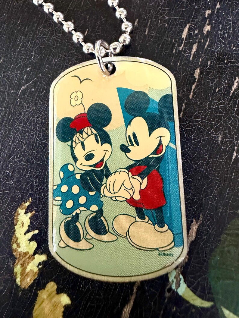 May include: A rectangular metal dog tag pendant on a silver ball chain. The pendant features a colorful graphic of Mickey and Minnie Mouse holding hands. Minnie wears a blue polka dot dress and red hat, while Mickey wears red shorts. The text Disney is visible.