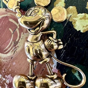 May include: A gold-coloured Mickey Mouse brooch in a cheerful pose, with one hand raised in a wave. The brooch has a shiny finish and detailed features, including Mickey's iconic ears and gloves. The pin is a collectible item.