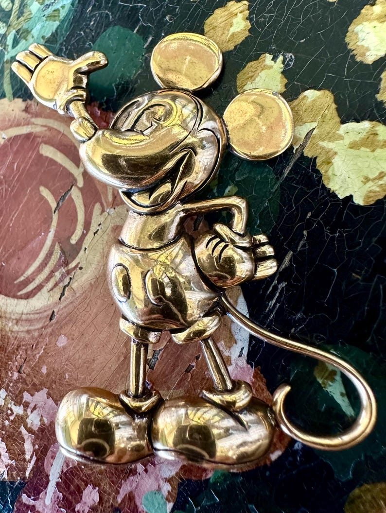 May include: A gold-coloured Mickey Mouse brooch in a cheerful pose, with one hand raised in a wave. The brooch has a shiny finish and detailed features, including Mickey's iconic ears and gloves. The pin is a collectible item.