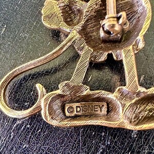 May include: A vintage gold-toned metal Disney pin depicting a stylized character. The pin has detailed features and a classic design. The word "DISNEY" is stamped on the base, indicating its origin.