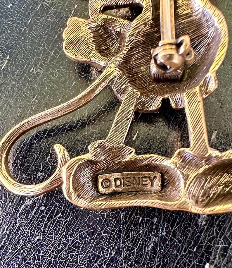 May include: A vintage gold-toned metal Disney pin depicting a stylized character. The pin has detailed features and a classic design. The word "DISNEY" is stamped on the base, indicating its origin.