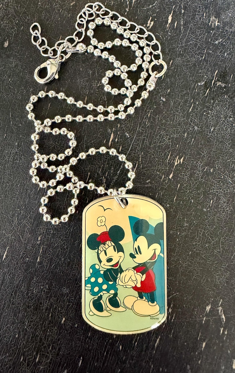 May include: A silver-toned ball chain necklace featuring a rectangular dog tag pendant. The pendant displays a colorful illustration of Mickey and Minnie Mouse holding hands, set against a light yellow background with a small bird silhouette above.