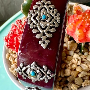 May include: A burgundy-colored bangle bracelet with ornate silver-tone metalwork and turquoise accents. The bracelet features a rectangular shape with decorative filigree designs and a glossy finish. The jewelry is displayed among small rocks and cacti.
