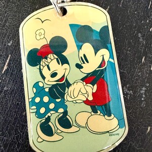 May include: A rectangular metal dog tag pendant on a silver ball chain featuring a colorful illustration of Mickey and Minnie Mouse holding hands. Minnie wears a blue polka dot dress and a red bow, while Mickey wears red shorts. The text Disney is visible.