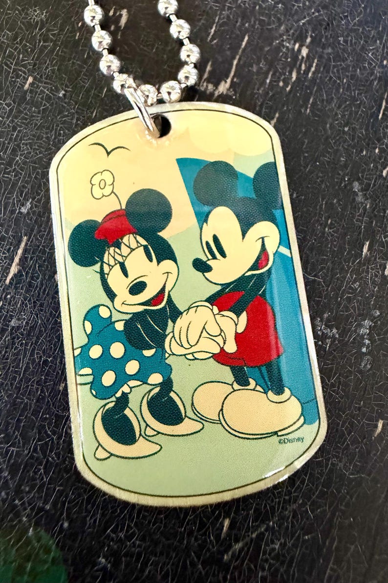 May include: A rectangular metal dog tag pendant on a silver ball chain featuring a colorful illustration of Mickey and Minnie Mouse holding hands. Minnie wears a blue polka dot dress and a red bow, while Mickey wears red shorts. The text Disney is visible.