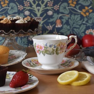 Peut inclure: Une tasse et une soucoupe blanches avec des motifs floraux, accompagnées de cupcakes, de fruits et de tranches de citron sur une table en bois. La tasse présente des fleurs roses et jaunes. Des cupcakes avec glaçage sont sur un présentoir à gâteaux en verre.