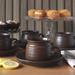 以下が含まれることがあります： 茶色のセラミックティーカップとソーサーのセット。カップはリブのデザインで、カップケーキとレモンスライスが置かれた木製のテーブルに置かれています。ガラスのケーキスタンドには、さらにカップケーキが置かれています。
